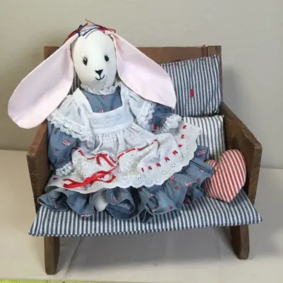 Handmade cloth rabbit doll dressed in a ruffled lace apron dress, seated on a miniature wooden bench with striped fabric cushions.