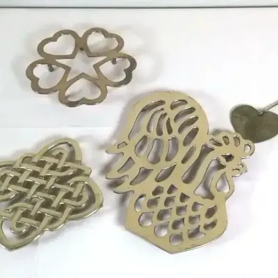 Assortment of brass metal tabletop decor including three trivets with rooster, knotwork, and heart patterns alongside a pair of...
