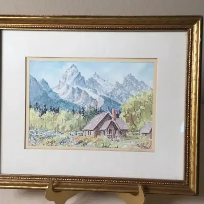 Framed mountain landscape painting depicting a chapel against snow-capped peaks, double-matted in a gilt wood frame.