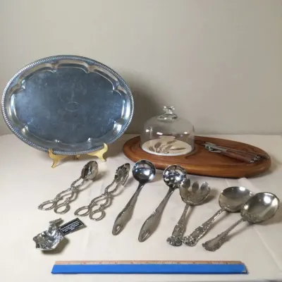 Collection of vintage silver-plated serving utensils and tableware, including a oval tray, salad servers, ladles, and a wooden cheese...