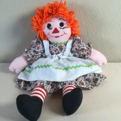 Handmade Raggedy Ann rag doll featuring bright orange yarn hair, a floral dress with a white apron, and classic red-and-white striped legs.