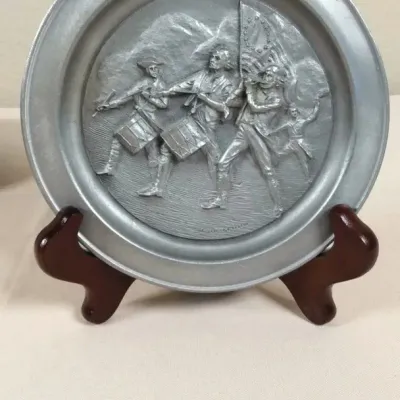Vintage pewter commemorative plate featuring a high-relief relief scene of "The Spirit of '76," displayed on a dark wooden...