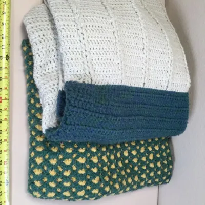 Handmade crocheted blankets folded in a stack, featuring a white and teal throw on top of a green and yellow patterned afghan.