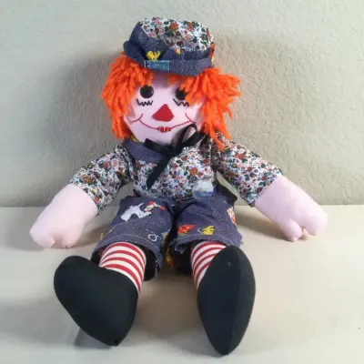 Handmade cloth Raggedy Andy-style doll featuring bright orange yarn hair, a floral print shirt, denim overalls, and red-and-white striped...