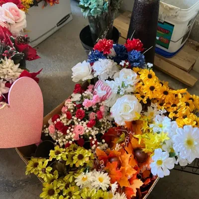Cardboard box overflowing with various colorful artificial floral stems, patriotic decorations, and autumn foliage.