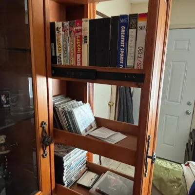 Wooden media storage cabinet featuring pull-out side shelves filled with VHS tapes, CDs, and DVDs.