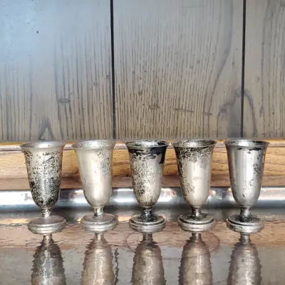 Set of five vintage silver-plated cordial glasses with tarnish and patina, displayed on a reflective surface.