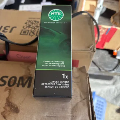 Boxed NTK oxygen sensor resting on cardboard shipping boxes in a workshop or storage area.