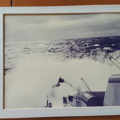 Framed black-and-white photograph depicting a naval ship deck cutting through rough ocean waves.