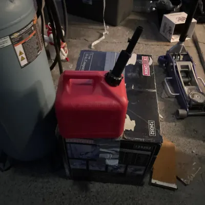 Red plastic gas can resting on a boxed Craftsman tool, alongside a vertical air compressor and a hydraulic floor jack in a garage setting.