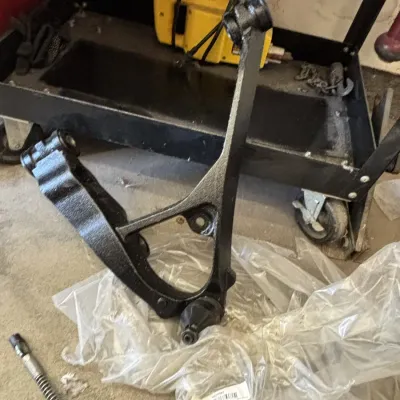 Black painted automotive control arm resting on plastic wrap near a utility cart and power tools.