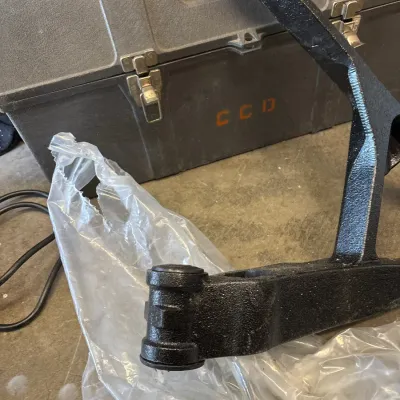 Black painted automotive suspension control arm resting on plastic wrapping in front of a gray molded plastic hard case.