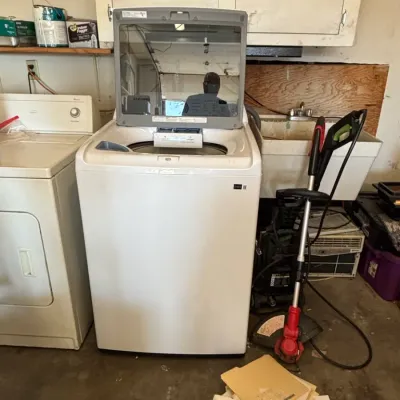 Modern white top-loading washing machine with a clear glass lid, sitting next to a vintage clothes dryer in a garage setting.