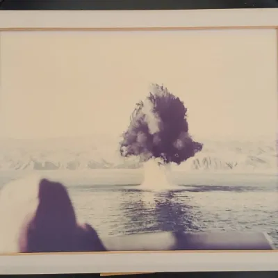 Framed vintage photograph depicting a mushroom cloud explosion over a body of water, set in a light wood frame with a wide mat.