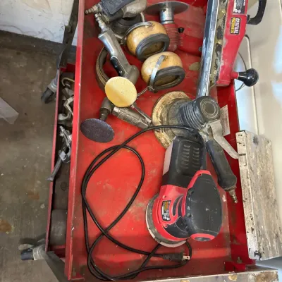 Red rolling tool cart filled with assorted pneumatic body shop tools, electric palm sanders, die grinders, and ear protection, showing...