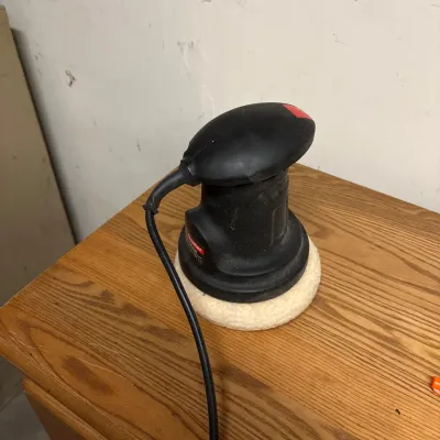 Handheld electric car buffer and polisher with a black plastic housing and a white buffing bonnet, resting on a wooden surface.