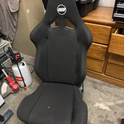 Black fabric bucket racing-style car seat featuring a white embroidered logo and harness passthrough cutouts.