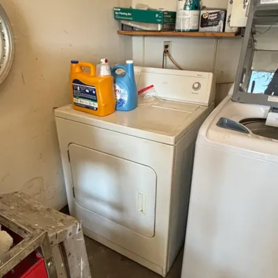 White front-loading Whirlpool dryer set in a utility room, accompanied by laundry detergent bottles resting on top.