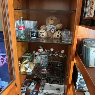 Wooden display cabinet filled with various knick-knacks, glassware, a plush teddy bear, holiday figurines, and media cases.