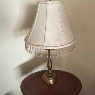 Traditional brass table lamp featuring a cream fabric lampshade trimmed with hanging crystal beaded fringe, set on a dark wood side table.