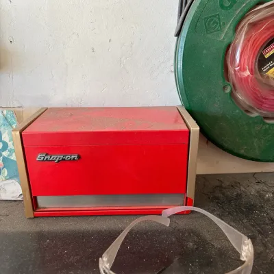 Vintage miniature red metal Snap-on jewelry or trinket chest, shown alongside clear plastic safety glasses and a Greenlee hose reel.
