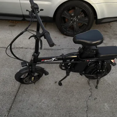 Compact black Sport e-bike featuring a folding frame, rear passenger seat, and disc brakes, parked on a concrete driveway.