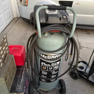 McGraw 21-gallon 175 PSI vertical portable air compressor with attached hose, shown in used condition alongside other driveway tools.