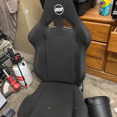 Black fabric racing-style bucket seat featuring integrated harness pass-through slots and printed headrest logo.