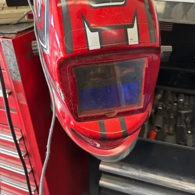 Auto-darkening welding helmet featuring a red shell with black and silver graphic accents, shown mounted near a red tool chest.