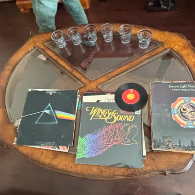 Collection of classic rock vinyl LP records, a 45 RPM single, and branded glassware displayed on a wooden glass-top coffee table.