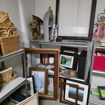 Sale Photo Thumbnail #58: Collection of framed artwork, empty picture frames, wicker baskets, and nautical-themed decor displayed on metal shelving units.