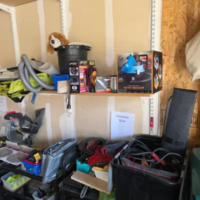 Sale Photo Thumbnail #146: Assorted garage and auto supplies including a boxed 12-volt wet/dry auto vacuum, miter saw, tarps, and jumper cables arranged on shelving and storage bins.