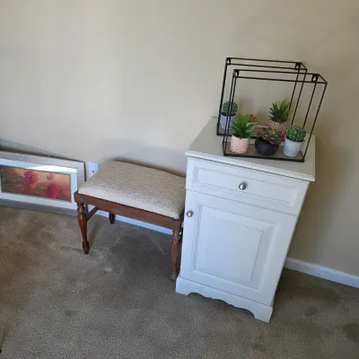 Sale Photo Thumbnail #94: White single-door nightstand with a top drawer alongside a wood-framed upholstered vanity bench.