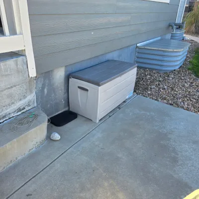 Sale Photo Thumbnail #164: Large beige plastic outdoor deck storage box with a dark grey hinged lid, situated on a concrete walkway against a house wall.