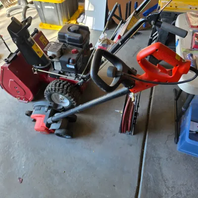 Sale Photo Thumbnail #129: Red gas-powered two-stage snow blower shown alongside an electric lawn edger and a hose reel in a garage setting.