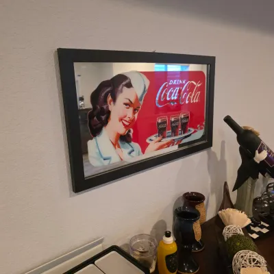 Sale Photo Thumbnail #10: Framed Coca-Cola mirror featuring a vintage-style waitress illustration, displayed above a tabletop with decorative items.