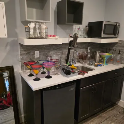 Sale Photo Thumbnail #28: Collection of colorful hand-blown glass margarita glasses and barware displayed on a wet bar countertop, alongside a mini fridge, microwave, and wine opener station.