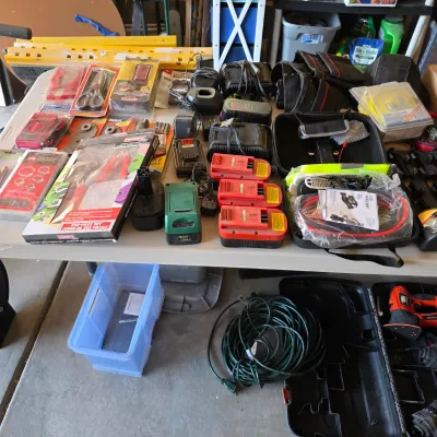 Sale Photo Thumbnail #128: Tabletop assortment of power tool batteries, chargers, hand tools, digital multimeter, and a coiled extension cord below.
