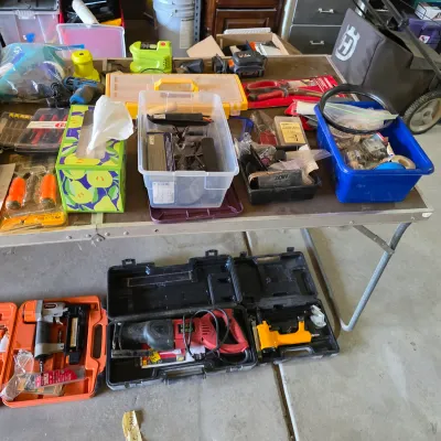 Sale Photo Thumbnail #125: Assortment of power tools, hand tools, and hardware organizers, including pneumatic nailers, a reciprocating saw, and various storage bins arranged on a folding table and floor.