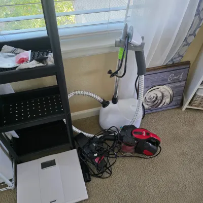 Sale Photo Thumbnail #92: Collection of small household appliances including a standing garment steamer, a handheld dustbuster vacuum, and a digital bathroom scale sitting near a black utility shelf.