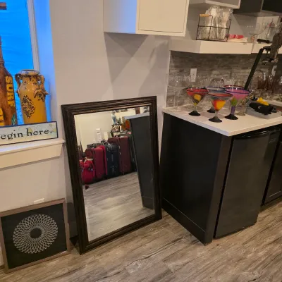 Sale Photo Thumbnail #27: Large dark wood framed floor mirror leaning against a wall, flanked by framed wall art and a kitchen counter set with colorful margarita glasses.