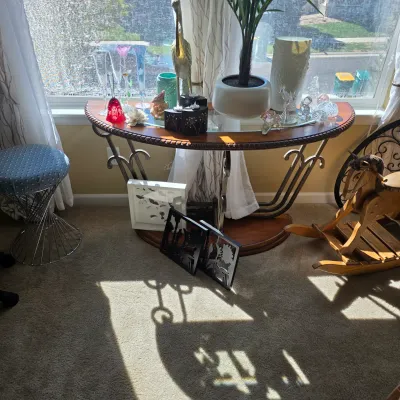 Sale Photo Thumbnail #99: Half-moon console table with wrought iron legs and glass insert top, surrounded by glassware, decor, a modern vanity stool, and a wooden rocking horse.