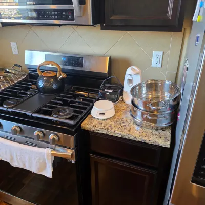 Sale Photo Thumbnail #114: Kitchen countertop with small appliances including a digital food scale, electric can opener, alarm clock, stacked stainless steel steamer pots, and a black hammered tea kettle on a gas range.