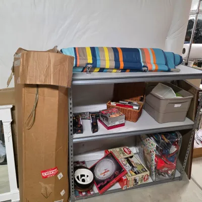 Sale Photo Thumbnail #51: Metal shelving unit filled with various household items, including a portable gas grill box, decorative serving trays, plastic storage bin, and a striped padded lounge mat.