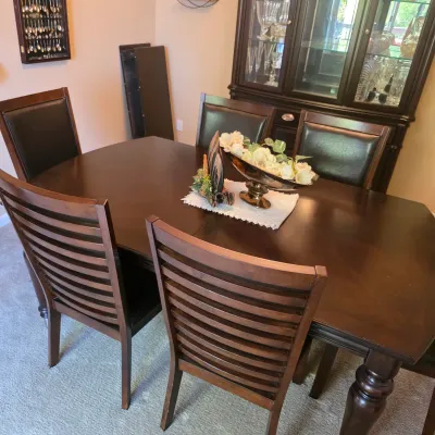 Sale Photo Thumbnail #76: Dark wood dining set including an rectangular expandable table and six matching chairs with slatted and upholstered backs, positioned in front of a glass-front china cabinet.
