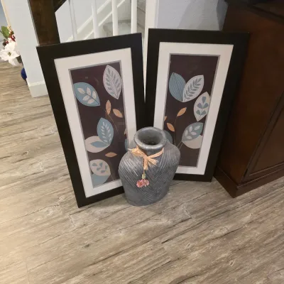 Sale Photo Thumbnail #5: Pair of framed botanical wall art prints featuring stylized leaf designs alongside a textured grey ceramic vase with decorative raffia ribbon.