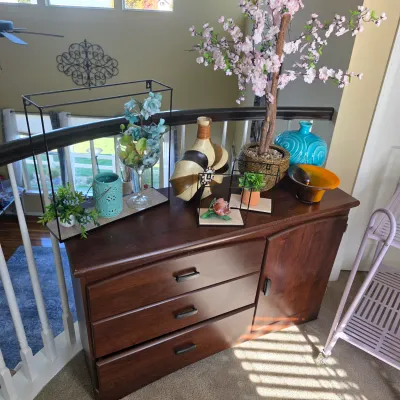 Sale Photo Thumbnail #85: Dark wood console cabinet with three drawers and a side cupboard door, styled with an assortment of home decor including a faux cherry blossom tree, wire frame displays, ceramic vases, and faux greenery.