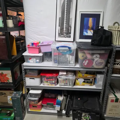 Sale Photo Thumbnail #63: Metal utility shelving unit packed with plastic storage totes, craft supplies, lightbulbs, and framed art prints including a black-and-white photograph of the Flatiron Building.