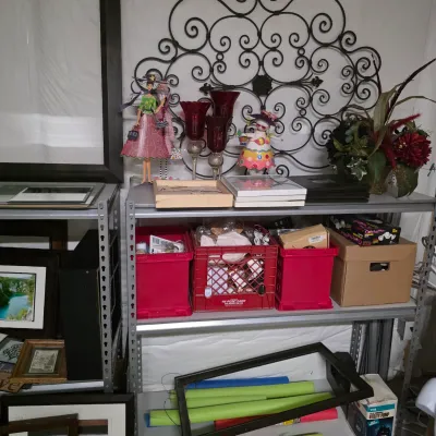 Sale Photo Thumbnail #57: Metal shelving unit holding decorative home accents, red glassware, figurines, storage bins, framed artwork, and pool noodles.