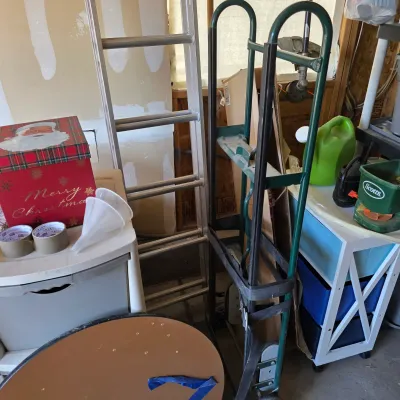 Sale Photo Thumbnail #134: Collection of garage and household items, including a green hand truck, an aluminum ladder, storage drawers, lawn care equipment, and holiday decorations.
